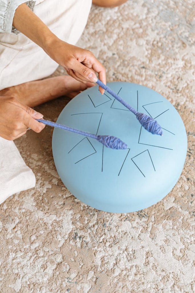 Close-up of hands playing a blue steel tongue drum with mallets on a textured carpet.